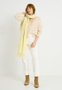 Cárdigan beige de punto con cierre de botones, combinado con una bufanda larga amarilla con bordes con flecos. Pantalones blancos remangados y botas tobilleras de color camel.