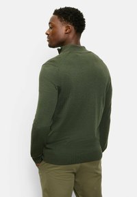 Green knit pullover with a high collar, textured fabric, and ribbed cuffs. Worn with olive-green pants, features seamless shoulder design.