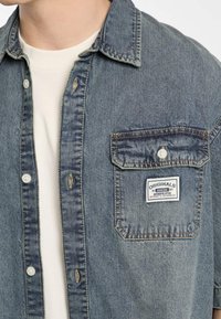 Denim overhemd met een lichtblauwe wassing, voorzien van een puntige kraag, knopenlijst aan de voorkant en een borstzak met een label. Gelaagd, casual uiterlijk.