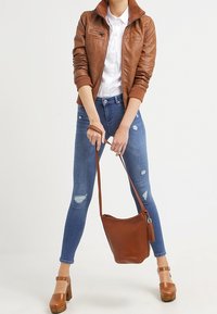 Giacca in pelle marrone con zip e polsini a coste, camicia bianca con bottoni, jeans skinny blu consumati e borsa a tracolla marrone. Scarpe platform color cannella.