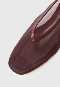 Scarpa bordeaux con punta affusolata, sovrapposta da rete trasparente e rifiniture in pelle, su sfondo bianco.