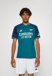 Maglia da calcio teal con accenti blu navy, dotata di colletto rotondo e maniche corte. Logo "Emirates Fly Better" in evidenza. Pantaloni corti bianchi.