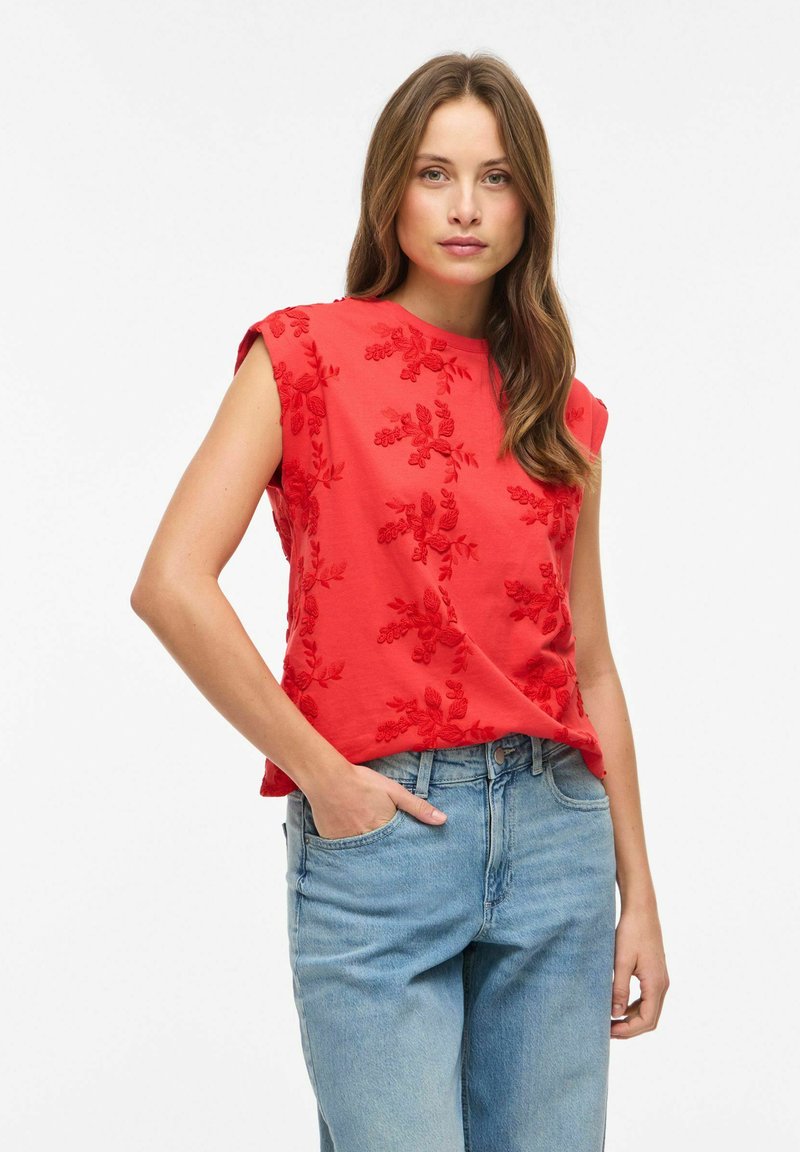 Nő piros ujjatlan, virágos hímzéssel díszített felsőben és kék magasderekú farmerral, egyik kezét zsebében tartva, fehér háttér előtt áll.