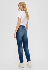 Jeansy o wysokim stanie, proste nogawki w średnim niebieskim denimu, z dwoma tylnymi kieszeniami i subtelnym pomarańczowym wykończeniem szwów. Noszone z białą koszulką.