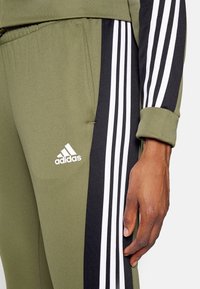 Gros plan d'un pantalon de survêtement Adidas vert olive avec des panneaux latéraux noirs et des bandes blanches, montrant une main reposant à côté du pantalon.