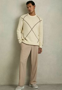 Krēmveida adīta džemperis ar brūnu rombu rakstu, pārī ar gaismas beigu platām biksēm un pelēkiem slip-on apaviem. Visiem apģērba elementiem ir gluda tekstūra.