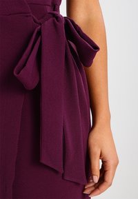 Tessuto di abito viola intenso con una finitura testurizzata, caratterizzato da una grande fascia annodata legata in vita, che crea un aspetto fluente.