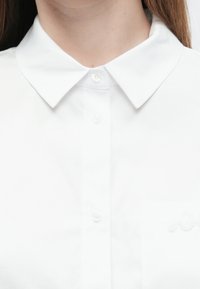 Valkoinen nappipaita klassisella kauluksella, jossa on sileä kangas, yksi rintatasku ja brodeerattu logotyyli.