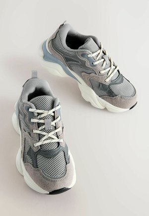 Sive teniske, ki združujejo mrežaste in semiš materiale, zaobljena oblika, kontrastna gumijasta podlaga, bele vezalke in teksturirani detajli.