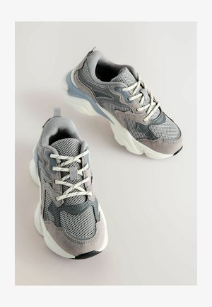 Grå sneakers med mesh- og ruskindmaterialer, rundet design, kontrasterende gummisål, hvide snørebånd og teksturerede accenter.