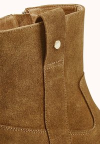 Bottine en daim marron avec un bout arrondi, des surpiqûres contrastantes et un détail à bouton pression sur le côté. Doublure intérieure lisse.