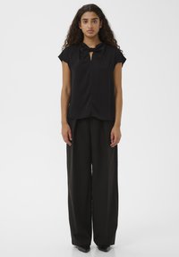 Donna con capelli ricci che indossa un top senza maniche nero con un collo a fiocco e pantaloni neri a gamba larga, in piedi davanti a uno sfondo semplice.