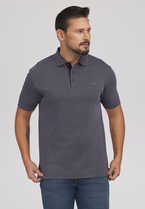 Mężczyzna z brodą, ubrany w ciemnoszarą koszulkę polo z krótkim rękawem i niebieskie dżinsy, stojący na tle jednolitego jasnego tła.