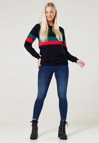 Schwarzer Pullover mit grünen und roten horizontalen Streifen, kombiniert mit blauen Skinny-Jeans und schwarzen Schnür-Stiefeletten. Glatte Textur.