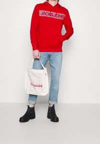 Sudadera roja con logo blanco, vaqueros claros de tiro alto arremangados, botas chelsea negras y una bolsa de mano blanca con "CAMBIO" impreso en gris y rojo.