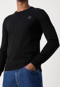 Czarny sweter z długim rękawem z prążkowanymi mankietami i pasem, wykonany z lekkiej dzianiny, z małą naszywką z logo na piersi. Połączony z niebieskimi dżinsami.