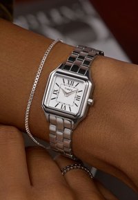 Sidabro spalvos nerūdijančio plieno laikrodis su kvadratiniu ciferblatu, baltu ciferblatu, juodais romėniškais skaitmenimis ir grandinėlės apyranke, užsegta ant įdegusios rankos.