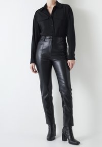 Pantaloni neri in pelle a vita alta con dettagli delle cuciture frontali, abbinati a una maglietta nera a maniche lunghe con due tasche sul petto, indossati da un modello.