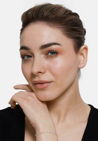 Nahaufnahme einer Person mit heller Haut, hellbraunen Haaren, die ein schwarzes Oberteil und ein zartes Silberarmband mit kleinen Steinen trägt. Blaue Augen.