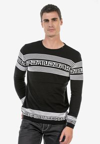 Schwarzer Pullover mit langen Ärmeln, der weiße horizontale Streifen, ein geometrisches Muster sowie ein geripptes Halsbündchen und Bündchen hat. Aus einem weichen Material gefertigt.
