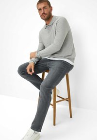 Grijze lange mouwen polo met drie knopen, gecombineerd met donkergrijze rechte jeans en witte sneakers, zittend op een houten kruk.
