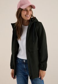 Veste légère vert foncé avec fermeture éclair, capuche réglable et poignets élastiques. Portée sur une chemise blanche et un jean bleu. Casquette en velours côtelé rose.