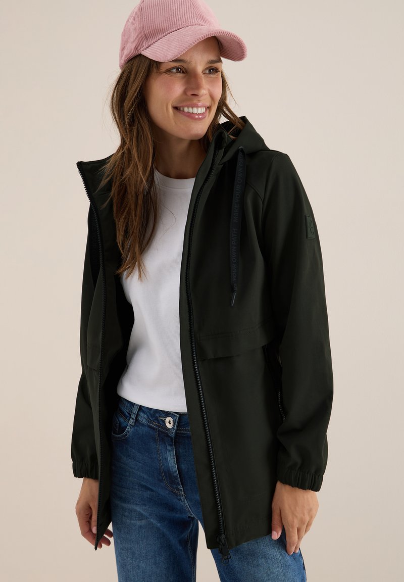 Veste légère vert foncé avec fermeture éclair, capuche réglable et poignets élastiques. Portée sur une chemise blanche et un jean bleu. Casquette en velours côtelé rose.