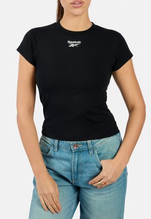 Vrouw draagt een aansluitend zwart Reebok t-shirt en lichtblauwe spijkerbroek met één hand in de zak en de andere rustend op de dij.