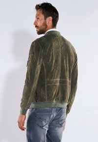 Braune Wildleder-Bomberjacke mit gerippten Bündchen und Saum. Verfügt über seitliche Reißverschlusstaschen und eine glatte Textur. Getragen mit blauen Jeans.