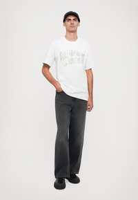 Tricou alb cu mânecă scurtă, cu imprimeu grafic, asortat cu blugi negri largi din denim și adidași negri. Modelul poartă de asemenea o șapcă neagră.