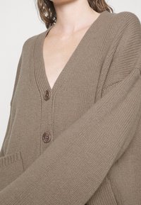 Frau trägt eine taubene V-Ausschnitt Strickjacke mit zwei großen braunen Knöpfen und strukturierten Ärmeln, die auf Brust- und Schulterhöhe gekürzt ist.