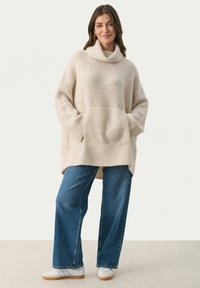 Beige strikket sweater med høj krave, oversized pasform, ribbet tekstur, to forlommer. Kombineret med bredbenede blå jeans og hvide sneakers.