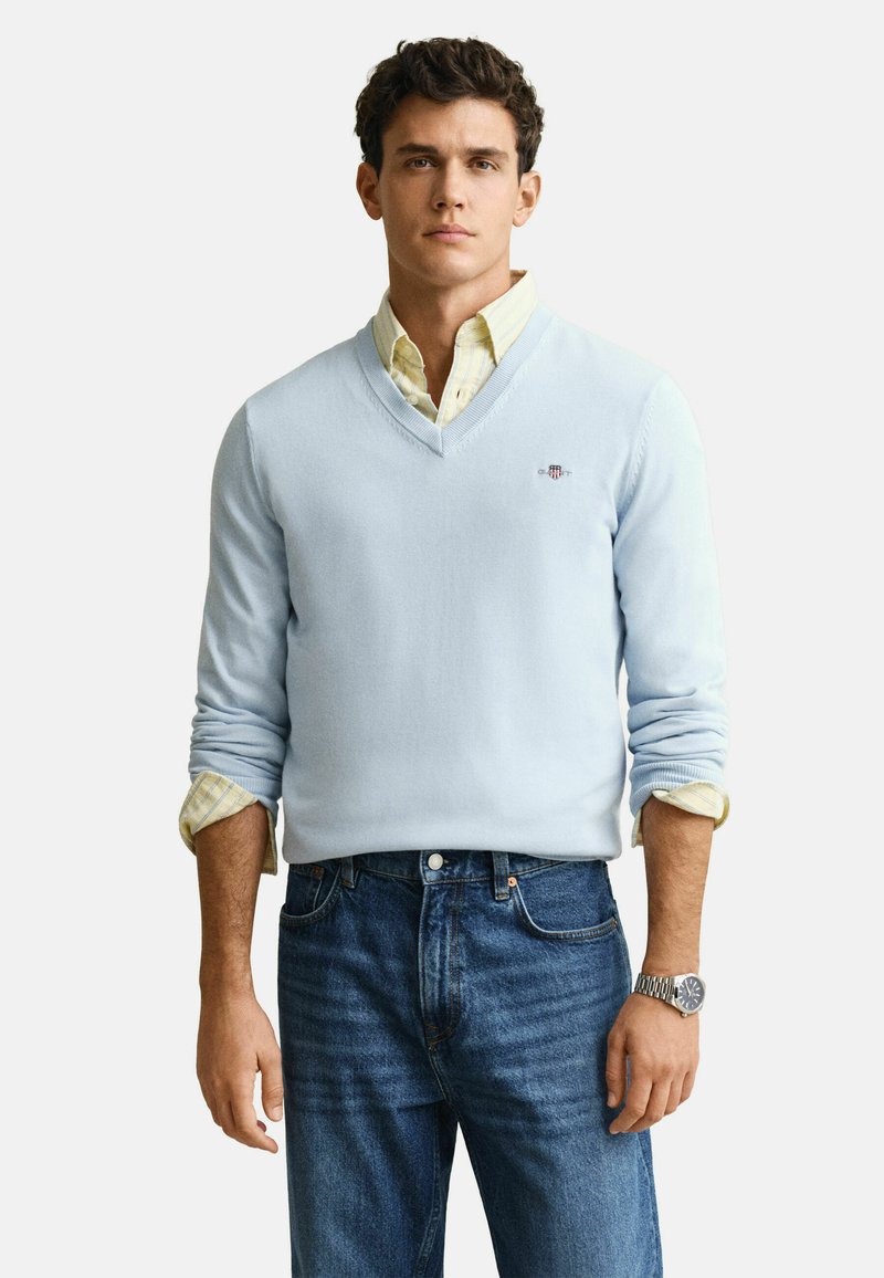 Homme portant un pull bleu clair à col en V par-dessus une chemise jaune rayée avec les manches retroussées, un jean bleu et une montre-bracelet argentée.