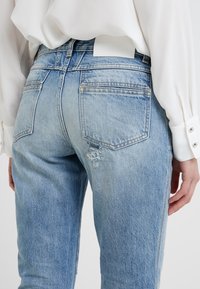 Jeans de mezclilla azul claro con un corte relajado, que presentan un detalle desgastado en la parte trasera. Llevados con una camisa blanca de manga larga.