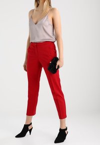 Donna che indossa un top in satin argento, pantaloni alla caviglia rossi sartoriali, stivaletti neri in suede con tacco, mentre tiene una piccola borsa a tracolla nera.
