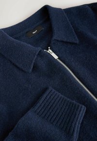 Granatowy sweter na zamek wykonany z miękkiego materiału, z kołnierzykiem, prążkowanymi mankietami i srebrnym detalem zamka. Etykieta widoczna w środku.
