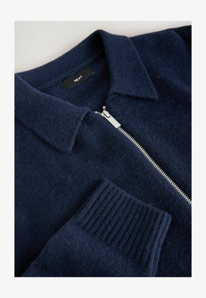 Granatowy sweter na zamek wykonany z miękkiego materiału, z kołnierzykiem, prążkowanymi mankietami i srebrnym detalem zamka. Etykieta widoczna w środku.