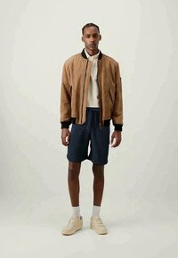 Brun mocka bomberjacka med svart krage, i kombination med marinblå shorts och beige sneakers. Minimalistisk design, mjuk textur och avslappnad passform.
