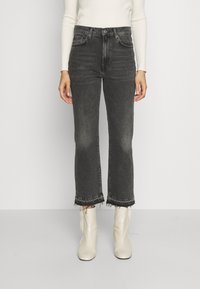 Persona que lleva unos pantalones vaqueros negros de tiro alto y cortos con dobladillos sin rematar, una camiseta de manga larga blanca de canalé y botas blancas hasta el tobillo, de pie en posición recta.