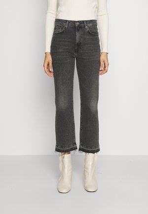 Persoon in hoge, zwarte cropped jeans met ruwe zomen, een wit geribbeld longsleeve shirt, en witte enkellaarsjes, rechtopstaand.