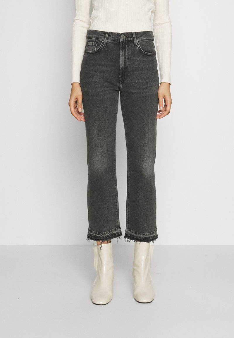 Persona que lleva unos pantalones vaqueros negros de tiro alto y cortos con dobladillos sin rematar, una camiseta de manga larga blanca de canalé y botas blancas hasta el tobillo, de pie en posición recta.
