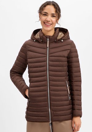 Smiling woman wearing a brown quilted jacket with a hood and zipper pockets, standing with one hand in pocket against a white background.