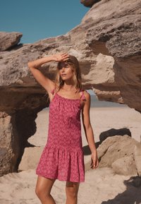 Abito estivo rosa con motivo a fantasia, spalline sottili, orlo sfrangiato e dettagli con nappine, su uno sfondo di spiaggia rocciosa.