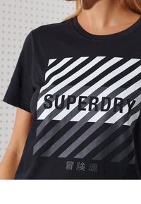 Sort bomulds T-shirt med et diagonalt stribet design i hvid og grå, med teksten "SUPERDRY" tydeligt vist.