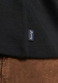 Schwarzer Rippstoff mit einem sichtbaren blauen "Superdry"-Etikett. Der Saum des Kleidungsstücks ist mit strukturierter Naht versehen, darunter befindet sich brauner Stoff.