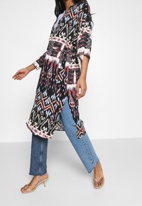 Long kimono imprimé aux couleurs sombres avec des motifs géométriques ; tissu léger, côtés fendus, associé à un jean bleu et des talons beiges à brides.