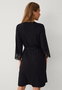 Robe noire avec des manches en dentelle trois-quarts, taille ceinturée et texture douce. Présente des détails en dentelle sur les bords des manches pour une touche élégante.