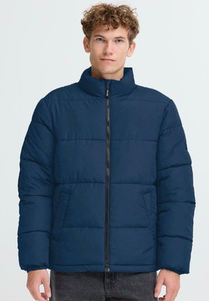 Veste matelassée marine avec col montant, motif matelassé horizontal, fermeture éclair sur le devant et poches latérales. Texture douce et lisse.