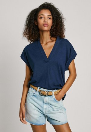 Maglietta blu navy a V con maniche corte arrotolate, abbinata a pantaloncini di jeans blu chiaro e una cintura intrecciata beige. Stile casual, vestibilità rilassata.