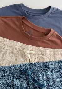 Tre beklædningsgenstande: en blå t-shirt, en rustfarvet t-shirt og mønstrede shorts med snøring. Teksturerede stoffer og varierede mønstre synlige.